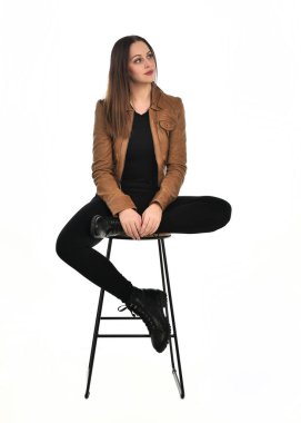 Kahverengi deri ceketli, modern siyah kıyafetler giyen güzel esmer kadın modelin tam boy portresi. Beyaz stüdyo arka planında izole bir figür..