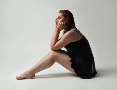 Güzel kadın modelin tam boy portresi, ıslak dağınık saçlar, sade siyah ipek bornoz, çürük bacaklar. Otururken duygusal hüzünlü bir poz verin. Stüdyo arka planında izole bir figür, koyu renkli karşıtlık gölgeleri.