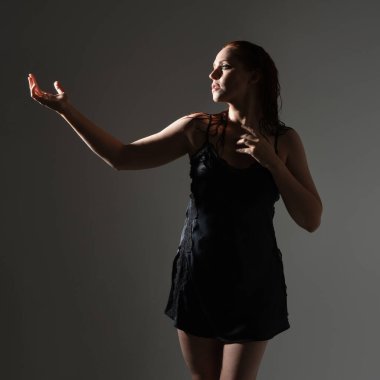 Kız modelin yakın portresi, ıslak dağınık saçlar, basit siyah ipek sabahlık, ayakta duran yan profil, gergin duygusal duruş, el hareketi. Karanlık stüdyo arka planında gölgeleri olan izole bir figür..