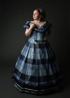 Viktorya dönemi kadınının tarihi klasik mavi balo elbisesi içinde tam boy portresi, güzel artı beden modeli. Karanlık stüdyo arka planında izole edilmiş siluet figürü.