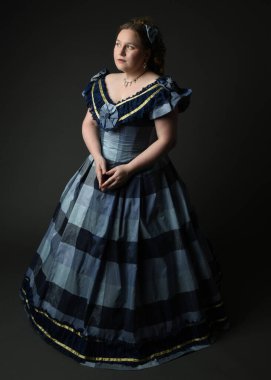 Viktorya dönemi kadınının tarihi klasik mavi balo elbisesi içinde tam boy portresi, güzel artı beden modeli. Karanlık stüdyo arka planında izole edilmiş siluet figürü.