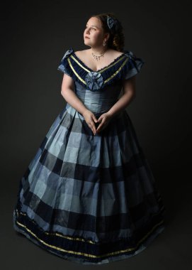 Viktorya dönemi kadınının tarihi klasik mavi balo elbisesi içinde tam boy portresi, güzel artı beden modeli. Karanlık stüdyo arka planında izole edilmiş siluet figürü.