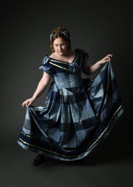 Viktorya dönemi kadınının tarihi klasik mavi balo elbisesi içinde tam boy portresi, güzel artı beden modeli. Karanlık stüdyo arka planında izole edilmiş siluet figürü.