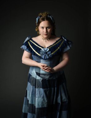 Viktorya dönemi kadınının tarihi klasik mavi balo elbisesi içindeki portresi, güzel artı beden modeli. Karanlık stüdyo arka planında izole edilmiş siluet figürü.