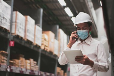 Maske takan Asyalı yakışıklı mühendis veya teknisyen, dijital tablet kontrol ürünleri stoku, güvenlik kargo yönetimi, depo, endüstri lojistik ve ihracat konsepti kullanıyor.