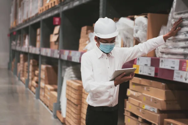 Asian handsome engineer or technician wearing mask,and safety hard hat,uses digital tablet check merchandise stock,security cargo management,in warehouse,industry business  logistic and export concept