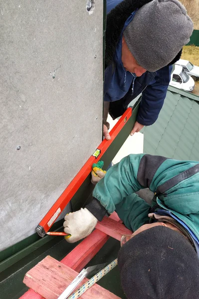 Workers line the chimney with green metal, work without insurance at height, use a level, tape measure and wooden scaffolding.