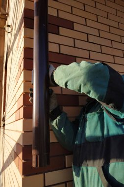 Ön tuğlayla örülmüş eve, işçi drenaj için boruları bağlıyor. Fotoğrafta duvar, kahverengi borular ve işçinin elleri görünüyor..