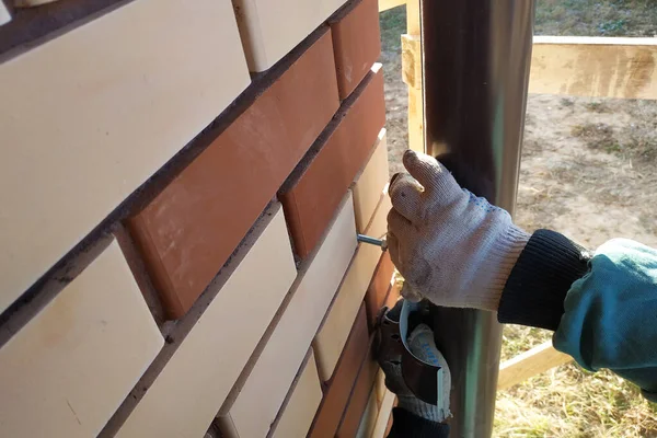 To the house bricked with a front brick, the worker fastens pipes for drainage. the photo shows the wall, brown pipes and the worker\'s hands 2020.
