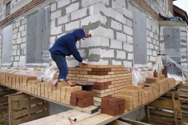 İşçi, evin duvarlarını kırmızı tuğlalarla örüyor. Yeni bir ev inşa etme süreci.