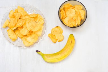 Açık bir tabağa yerleştirilmiş patates cipsleri ve olgun bir muzla birlikte bir kasenin içinde, hepsi beyaz ahşap bir masaya yerleştirilmiş.. 