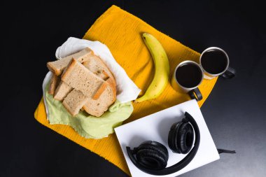 Kahvaltı malzemelerinin düz yatışı, dilimlenmiş ekmek, bir muz, iki fincan kahve, bir kitabın üzerinde dinlenen kulaklıklar, hepsi koyu bir arka planda sarı bir plasemat üzerine dizilmiş..