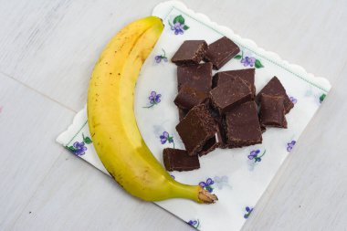 Olgun bir muz ve koyu çikolata parçaları hafif ahşap bir yüzey üzerindeki dekoratif bir peçetenin üzerinde dinleniyor. Basit ama çekici bir atıştırmalık ya da ikram öneriyor..