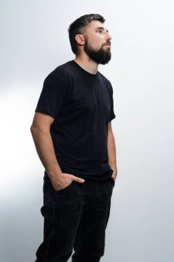Sıradan giyinmiş sakallı bir adam açık gri bir arka plana karşı duruyor. Koyu renk bir gömlek ve kot pantolon giyiyor, ellerini cebine sokuyor. Düşünceli bir şekilde kameradan dışarı bakıyor..