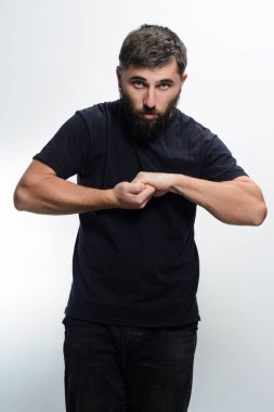 Koyu renk gömlekli ve kot pantolonlu sakallı bir adam beyaz arka planda duruyor ve eklemlerini kırıyor. Onun ciddi ifadesi ve doğrudan bakışı bir beklenti ya da gerilim hissi yaratır..