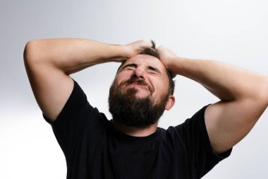 Sakallı bir adam kafasını iki eliyle kavrar, yüzü sıkıntı ve ıstırap ifadesiyle hafif bir arka plana çarpar. Güçlü duygularla boğulmuş gibi görünüyor..