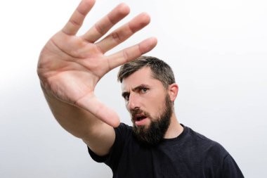 Beyaz arka planda sakallı bir adam elini kaldırır. Savunma ya da 'dur' işareti yapar. Yüzündeki ifade endişe ya da endişe gösterir..