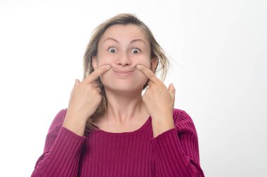 Beyaz arka planda kestane rengi süveter giyen bir kadın parmaklarını ağzının kenarlarını gülümsemeye zorlamak için kullanır..