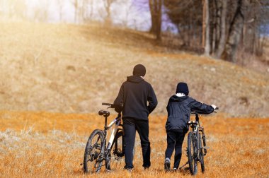 Bisikletleriyle yürüyen iki kişinin sakin bir sahnesi, altın, kuru otların ve parlak bir gökyüzünün altındaki ağaçlardan oluşan bir zemin üzerine kurulmuş. Barışçıl atmosfer aşikar..