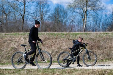 Genç bir çocuk ve ağabeyi toprak yolda bisiklet sürüyorlar. Güneş ışığı çimenli bir arazide akar. Bahar havası için sıkı giyinmişler..