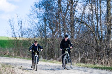 Toprak yolda bisiklet süren iki genç çocuk. Çıplak ağaçlar, bahar ya da sonbaharı işaret ediyor. Çocuklar siyah ceketler giydiler..