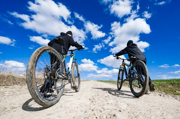 Tozlu bir patika boyunca dağ bisikleti süren iki kişinin düşük açılı çekimi. Açık gökyüzü tüylü beyaz bulutlar içerir, bisiklet maceraları için manzaralı bir zemin oluşturur.
