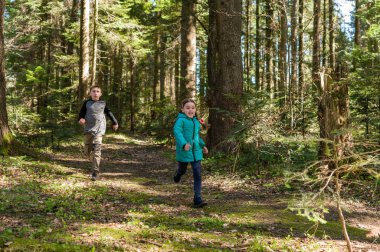 Bir çift kardeş, yemyeşil bir ormanda eğlenceli bir koşunun tadını çıkarıyor. Güneş ışığı ağaçların arasından süzülür, önlerindeki yolu aydınlatır..