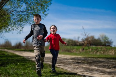 Gölgeli bir yolda koşan iki çocuğun mutlu sahnesi. Bereketli yeşil çimenler ve ağaçlar neşeli bir açık hava ortamı yaratıyor..