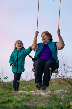 Dışarıda salıncaktan hoşlanan yaşlı bir kadın ve genç bir kızın yürek ısıtan bir sahnesi. Batan güneş sıcak bir parıltı saçar, neşe ve beraberlik hissini güçlendirir..