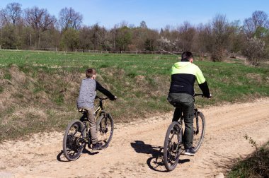 Bir adam ve bir çocuk toprak yolda bisiklet sürüyor, güneşli bir günün tadını çıkarıyorlar. Ağaçlar ve tarlalar onları çevreleyip barışçıl bir açık hava ortamı oluşturuyor. Sahne, aile bağlarını ve açık hava eğlencelerini çağrıştırıyor..