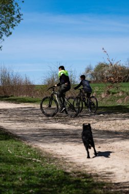 Açık mavi bir gökyüzünün altında toprak bir yol boyunca bisiklet süren iki insanın manzarası. Arkalarından bir köpek koşuyor. Ortam huzurlu ve güneşli..