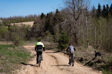 İki bisikletçi, muhtemelen baba oğul toprak yolda bisiklet sürüyorlar. Ağaçlar ve bir tarla parlak, güneşli bir gökyüzünün altında manzaralı bir zemin oluşturur. Yol kenarında bir köpek görünüyor..