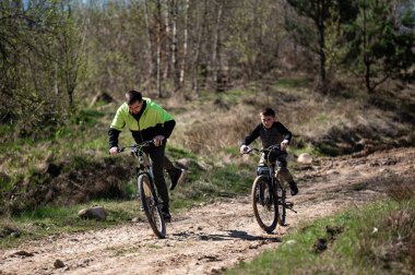 Bir adam ve bir çocuk ağaçlık bir alanda toprak bir patika boyunca bisiklet sürüyorlar. Güneş ışığı ağaçların arasından süzülüyor. Sahne bir aile eğlencesi ve açık hava macerası hissi uyandırıyor..