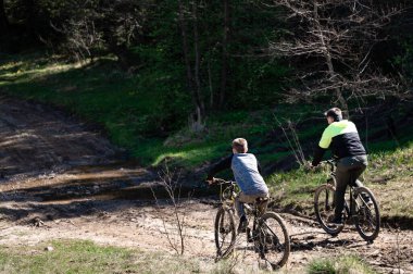 Dağ bisikletlerindeki iki figür toprak bir yolda ilerliyor. Bereketli yeşil yapraklar etraflarını sarar, güneşli ve açık hava maceralarını ima eder..