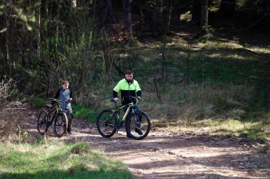 Bir adam ve bir çocuk ağaçlık bir alanda çamurlu bir yolda bisikletlerini durdurur. Güneş ışığı yolu karartır. Barışçıl bir aile aktivitesi.