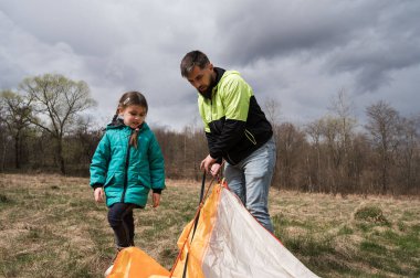 Bir baba kızına, yemyeşil bir orman ve dramatik gri gökyüzü ile çevrili renkli bir çadır kurarken rehberlik eder..