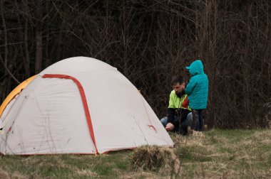 Parlak yeşil ceketli bir çocuk, narin dallar ve yumuşak çimlerle çevrili sakin bir orman açıklığında çadır kurarken bir yetişkine yardım eder..