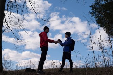 İki genç insanın siluetli figürleri doğal ortamda bir nesneyi değiş tokuş ediyorlar. Gökyüzü parlak mavi, kabarık bulutlar ve etrafını saran çıplak ağaçlar var..