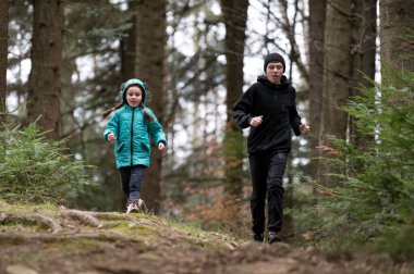 İki çocuk, temiz hava ve neşe dolu serin bir günde uzun ağaçlar ve canlı yeşilliklerle çevrili dolambaçlı bir orman yolunda neşeyle yarışıyor..