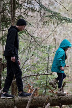 İki çocuk, renkli ceketlerle sarılı, sakin bir ormanda ağaçların oluşturduğu tuhaf bir atmosferle doğal çevreyi keşfederken ahşap bir patikadan geçiyor..