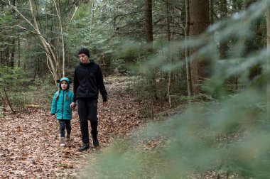 Gök mavisi ceketli bir çocuk, öğleden sonra macerasında yemyeşil ağaçlar ve renkli sonbahar yapraklarıyla çevrili huzurlu bir orman yolunda bir yetişkinin yanında yürüyor..