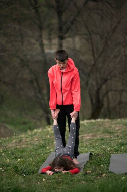 İki genç birey, yemyeşil bir ortamda, sakin bir öğleden sonra sıcak güneş ışığı altında takım çalışması ve dengeyi gösteren bir yoga pozisyonuna girerler..