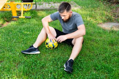 Sakallı bir adam, elinde sarı ve mavi bir futbol topuyla çimlerin üzerinde oturuyor. Bir maçtan ya da antrenmandan sonra ara vermiş gibi görünüyor. Arka planda bir fitness alanı görülebilir.