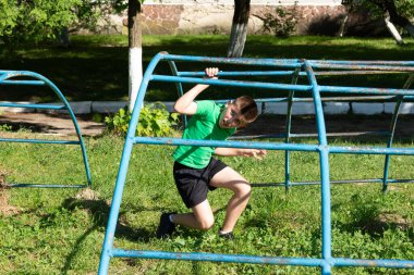 Genç bir çocuk çimenli bir parkta mavi bir metal tırmanış çerçevesiyle aktif olarak meşgul oluyor. Neşeli ve enerjik görünüyor, açık hava oyun ekipmanlarını seviyor..