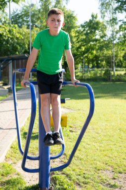 Genç bir çocuk açık havada paralel barlarda dalış yapıyor. Sahne yemyeşil otlarla dolu güneşli bir park. Spor kıyafetleri ve ayakkabıları var..