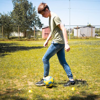 Genç bir adam çimenli bir alanda futbol antrenmanı yapıyor. Kot pantolon ve tişört giyiyor. Koniler çalışma alanını gösteriyor. Güneş parlak ve arka plan basit binaları gösteriyor..
