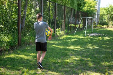 Genç bir insan çimenli bir arazide bir futbol topu taşıyarak yürür. Arka planda bir futbol golü görünür. Hava güneşli..