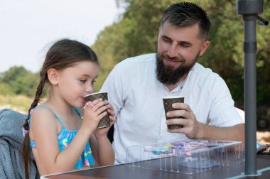 Sakallı bir adam ve küçük kızı açık bir masada oturup tek kullanımlık bardaklardan içmenin tadını çıkarıyorlar. Gülümsüyorlar ve mutlu görünüyorlar, keyifli bir günde birlikte güzel vakit geçiriyorlar..