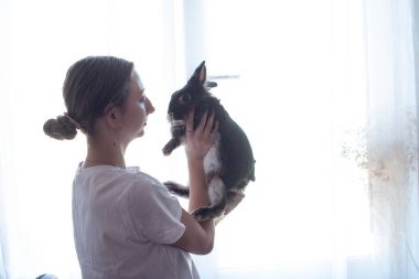 Genç bir kadın tüylü bir tavşanı kucaklıyor. Arkalarından gelen yumuşak bir ışık huzurlu ve huzurlu bir hava yaratıyor. Sahne samimi ve iç açıcı..