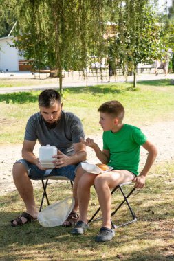Bir adam ve bir çocuk parkta portatif sandalyelerde oturur, paket servis konteynırlarındaki yiyecekleri paylaşırlar. Ortam sakin ve güneşli, bir ağacın gölgesinde..